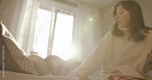 Wallpaper Mural A pleasant young girl holds a phone in her hands and calls her dog to jump upon the bed against the backdrop of sunlight from the window Torontodigital.ca