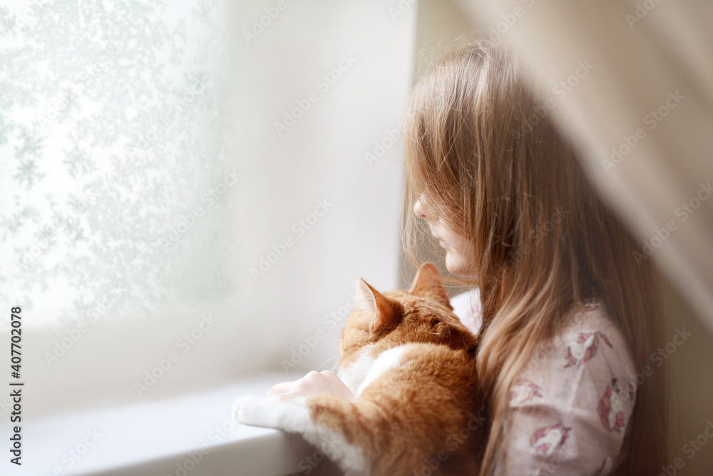 A child girl with a ginger cat in her arms looks out the window with frosty patterns. very soft artistic selective focus.