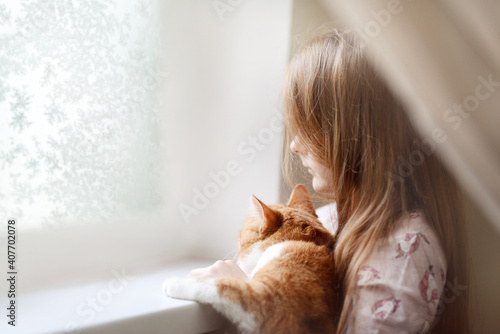 A child girl with a ginger cat in her arms looks out the window with frosty patterns. very soft artistic selective focus.