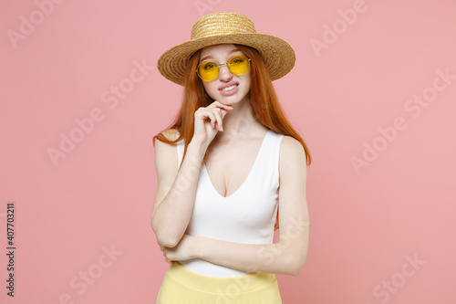 Young shy mistaken caucasian uncertain redhead woman 20s ginger long hair wear straw hat glasses summer clothes biting lip prop up chin look camera isolated on pastel pink background studio portrait