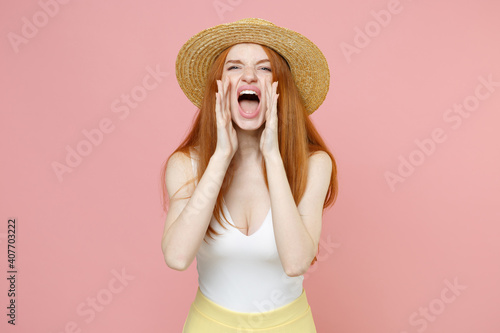 Young caucasian redhead woman 20s ginger long hair wearing straw hat and summer clothes shouting screaming news with hands arms near mouth isolated on pastel pink color background studio portrait