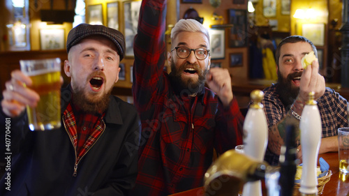 Cheerful friends having fun watching football game on tv and drinking draft beer at bar counter in pub.