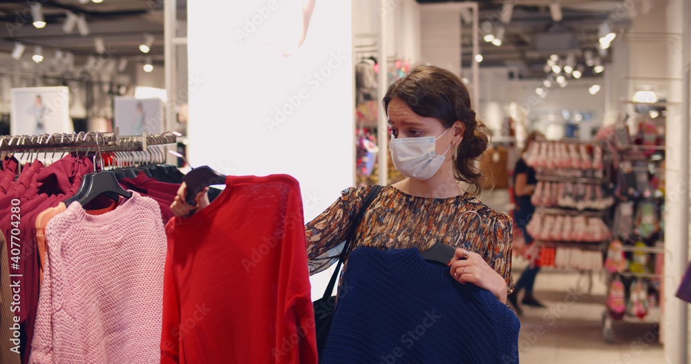 Young woman in medical mask choosing clothes in store