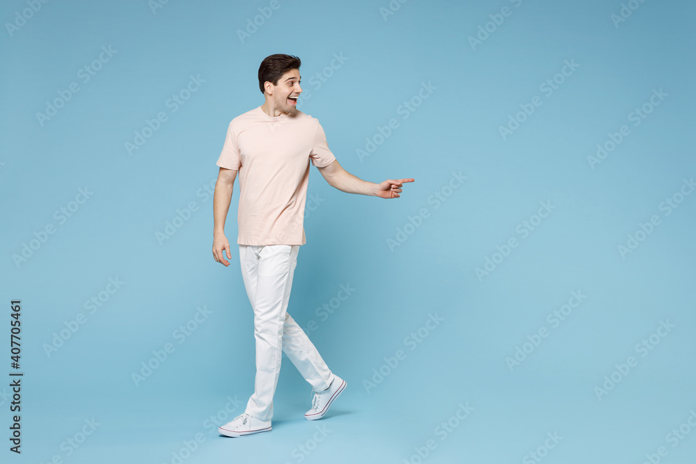 Naklejka premium Full length of young caucasian friendly smiling student man 20s in beige t-shirt white pants point index finger aside on copy space workspace area mock up isolated on blue background studio portrait.