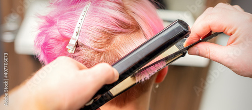 Close up of the hairdresser straightening the short pink hair of a female client with a hair straightening iron in a beauty salon