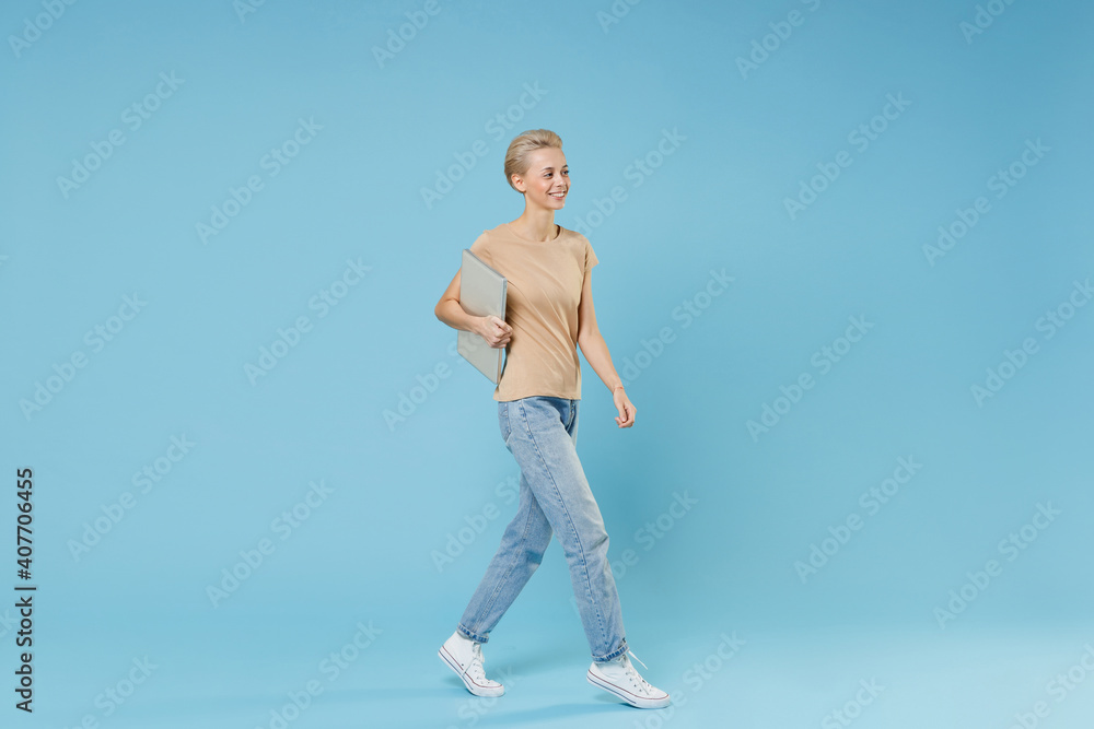 Full length of smiling caucasian young blonde woman 20s with short haircut wearing casual beige t-shirt jeans walking with closed laptop pc computer isolated on blue color background studio portrait. Full length of smiling caucasian young blonde woman 20s with short haircut wearing casual beige t-shirt jeans walking with closed laptop pc computer isolated on blue color background studio portrait.