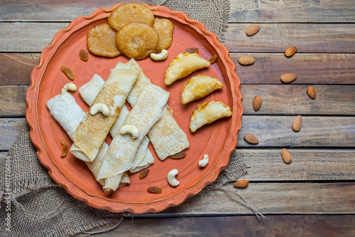 Bengali sweet - Patishapta Pitha, Malpua and Puli Pitha in a earthenware plate on wooden background with cashew nut, almonds and raisins.