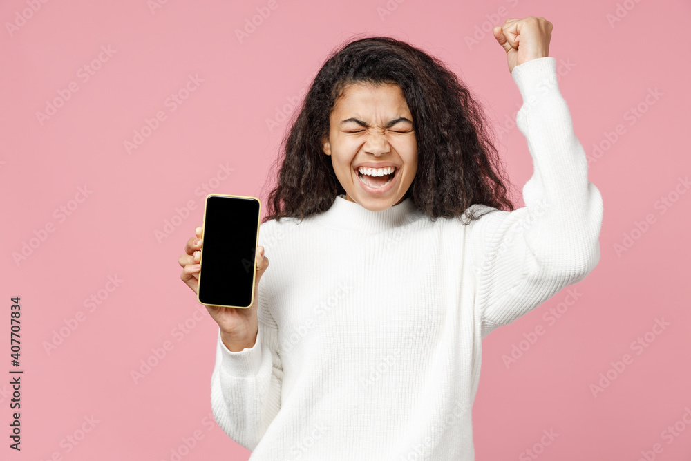 Young successful african american woman in white knitted sweater hold mobile phone with blank screen workspace area do winner gesture clench fist isolated on pastel pink background studio portrait Young successful african american woman in white knitted sweater hold mobile phone with blank screen workspace area do winner gesture clench fist isolated on pastel pink background studio portrait