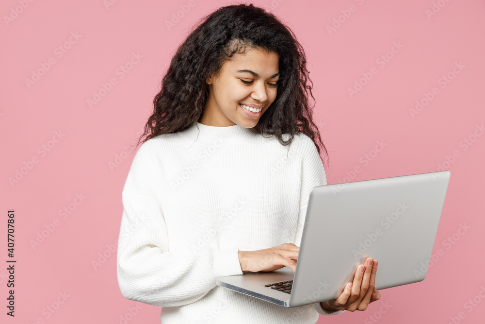 Young smiling cute african american woman 20s curly hair wearing white casual knitted sweater holding laptop pc computer typing chatting on keyboard isolated on pastel pink background studio portrait Young smiling cute african american woman 20s curly hair wearing white casual knitted sweater holding laptop pc computer typing chatting on keyboard isolated on pastel pink background studio portrait