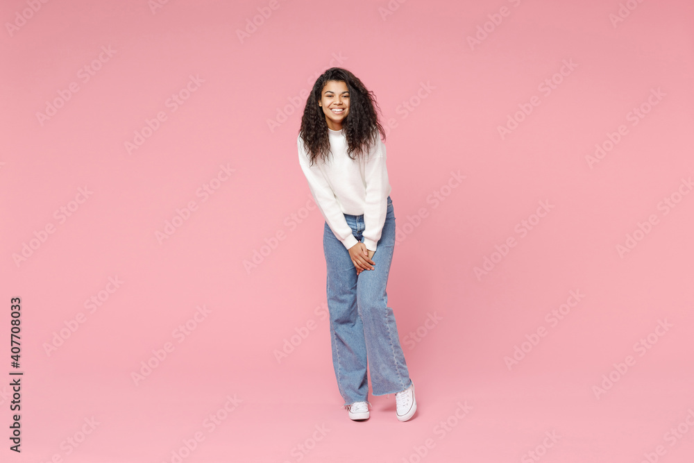 Full length young smiling cute african american woman 20s curly hair wear white casual knitted sweater jeans looking camera standing leaning over isolated on pastel pink background studio portrait. Full length young smiling cute african american woman 20s curly hair wear white casual knitted sweater jeans looking camera standing leaning over isolated on pastel pink background studio portrait.