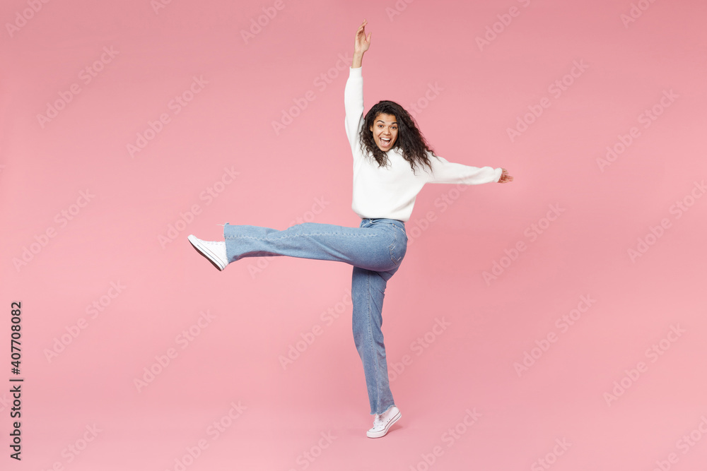 Full length young cute cheerful happy smiling african american woman 20s curly hair in white knitted sweater jeans raised up hand and leg fooling around isolated on pink background studio portrait. - obrazy, fototapety, plakaty 