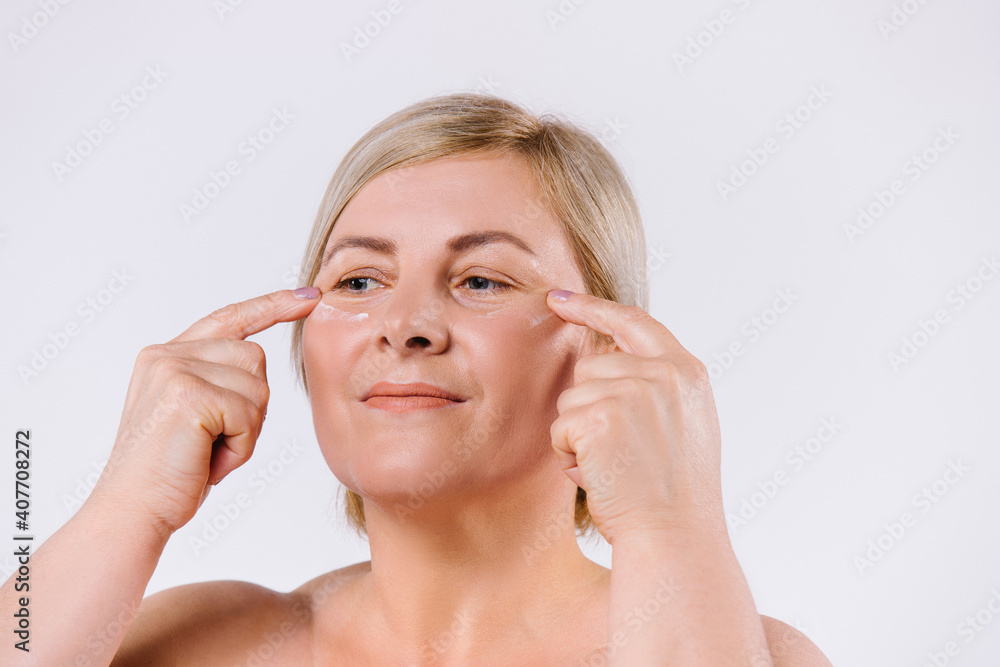 An older woman applies a revitalizing cream under her eyes with her index fingers. Enlarged photo. White background. An older woman applies a revitalizing cream under her eyes with her index fingers. Enlarged photo. White background.
