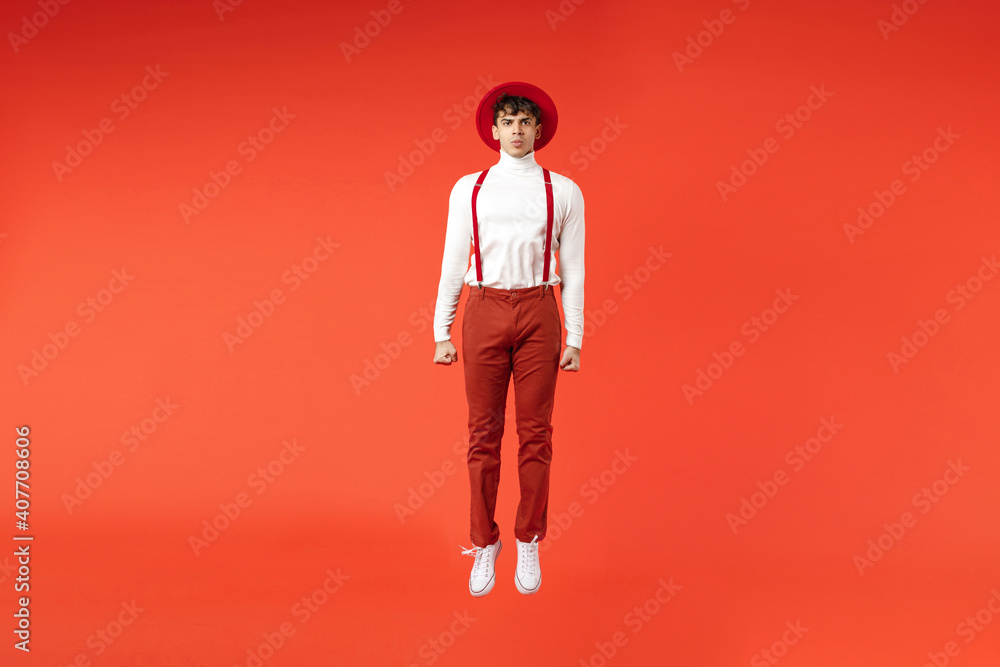 Full length of young spanish latinos serious cool fashionable stylish man 20s in hat white shirt trousers with suspenders jumping high looking camera isolated on red color background studio portrait. Full length of young spanish latinos serious cool fashionable stylish man 20s in hat white shirt trousers with suspenders jumping high looking camera isolated on red color background studio portrait.
