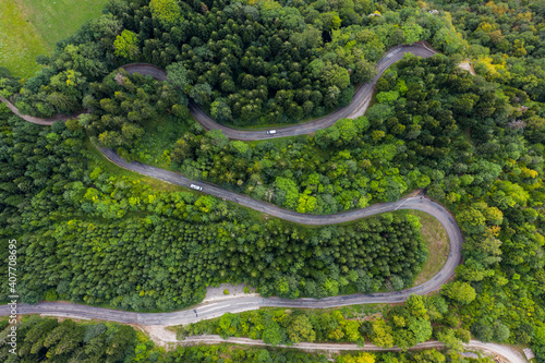 Aerial view of a winding countryside road passing through the green forest and mountain. White camping cars are passing through.