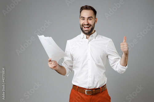 Excited young bearded business man wearing classic white shirt hold papers document showing thumb up like gesture isolated on grey color background studio portrait. Achievement career wealth concept.