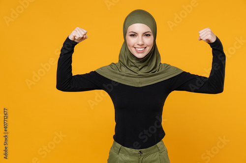 Smiling strong young arabian muslim woman in hijab black green clothes showing biceps muscles on hands isolated on bright yellow color background, studio portrait. People religious lifestyle concept.