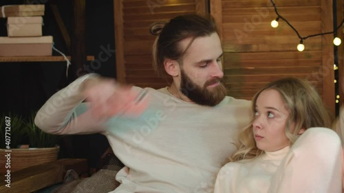 Young attractive couple is sitting on couch, watching TV. woman is on phone. Man takes the phone away from woman.