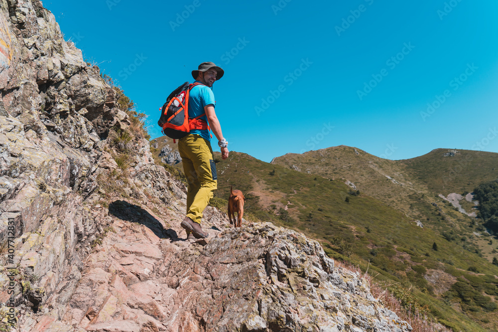 Happy young man hiker walking on rocky mountain ridge with his dog, rear view