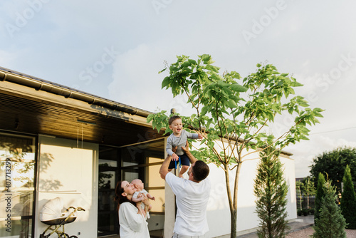 A young beautiful family with two little sons hug and kiss in the yard of their own house. Young mom and dad with two young sons hug and kiss in the yard of their own house.