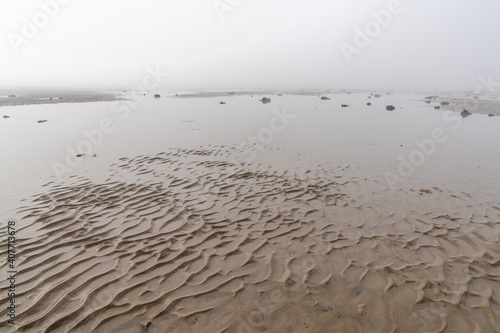 endless ocean floor uncovered at low tide with sand structures and rocks and tidal pools under lifting fog in the blue sky