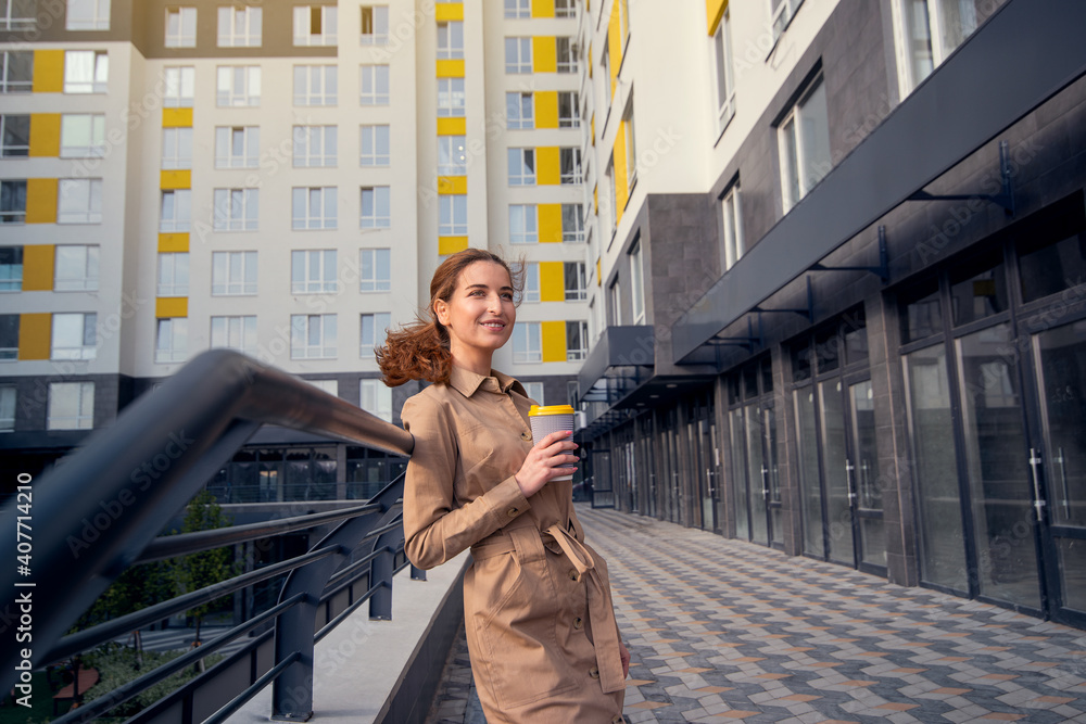 Young happy and successful woman wearing casual clothes holding a cup of coffee and standing against the background of high-rise building