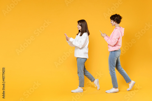 Full length side view of smiling two young women friends 20s wearing casual white pink hoodies using mobile cell phone typing sms message isolated on bright yellow color background studio portrait.