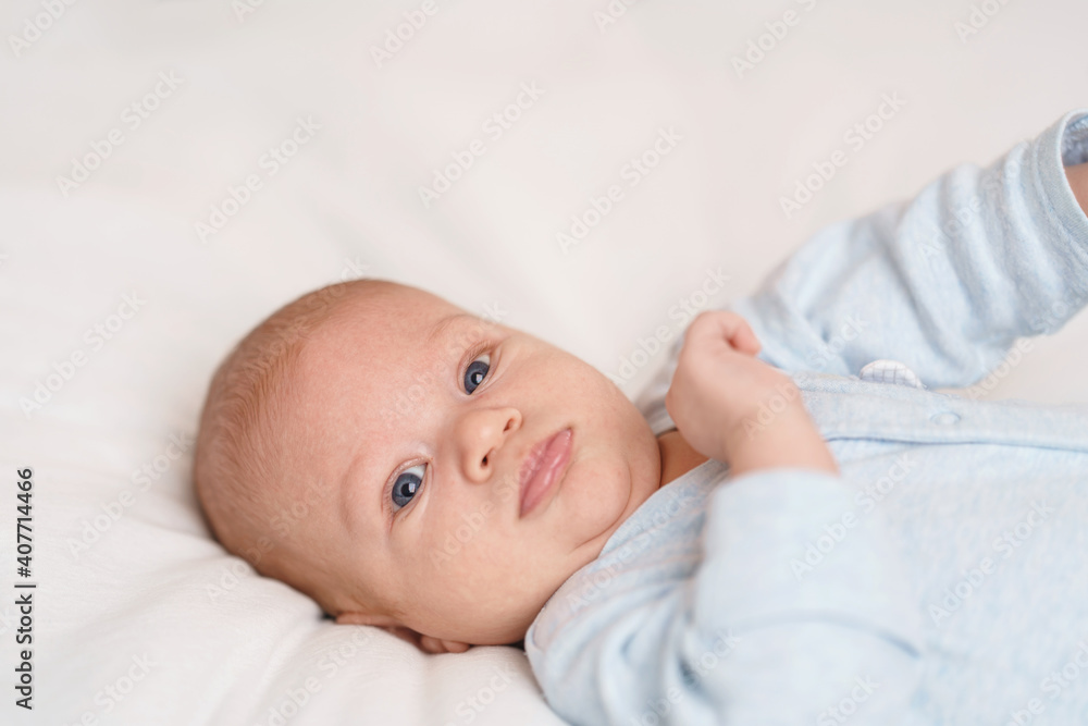 cute 2-month-old baby in blue bodysuit lies on bed and looks into frame cute 2-month-old baby in blue bodysuit lies on bed and looks into frame