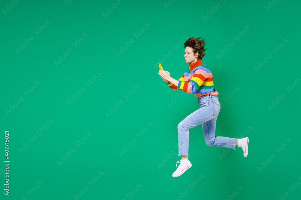 Full length side view of smiling funny cheerful young brunette woman 20s in colorful sweater jumping using mobile cell phone typing sms message isolated on green color background studio portrait.