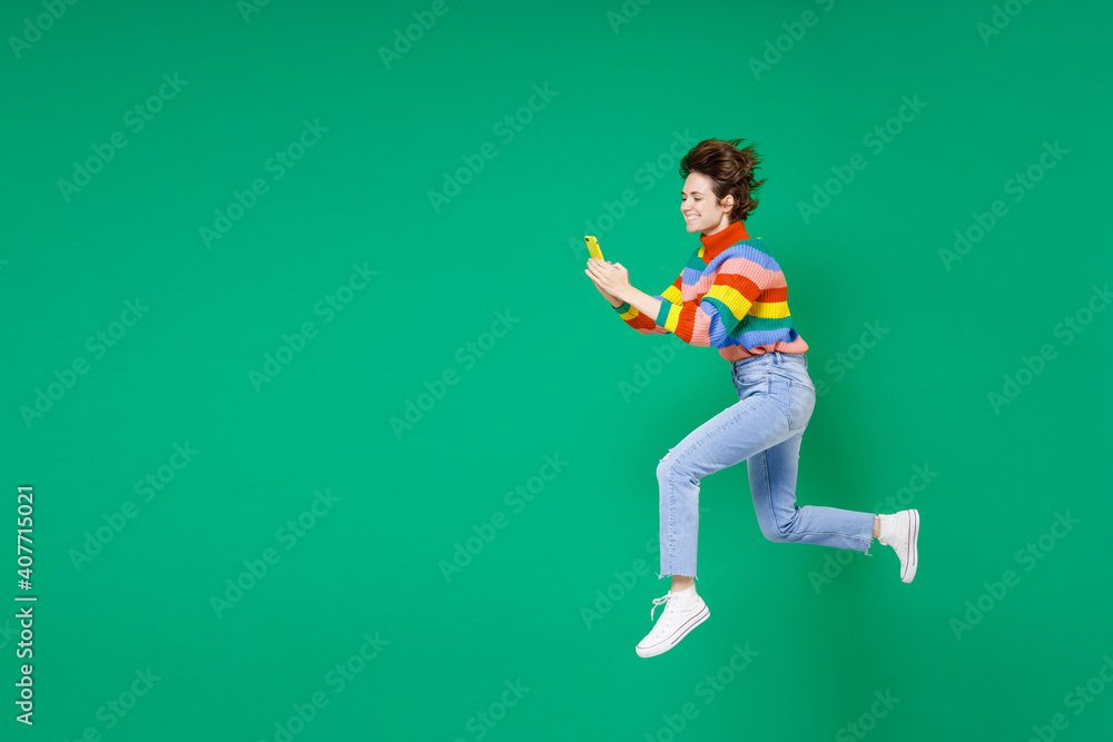 Full length side view of smiling funny cheerful young brunette woman 20s in colorful sweater jumping using mobile cell phone typing sms message isolated on green color background studio portrait. Full length side view of smiling funny cheerful young brunette woman 20s in colorful sweater jumping using mobile cell phone typing sms message isolated on green color background studio portrait.