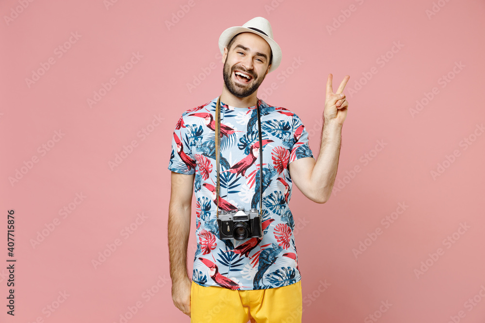 Naklejka premium Excited young traveler tourist man in summer clothes hat with photo camera showing victory sign isolated on pink background. Passenger traveling abroad on weekends getaway. Air flight journey concept.