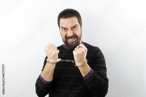 Portrait of an angry bearded man wearing casual clothes with handcuffs on white background