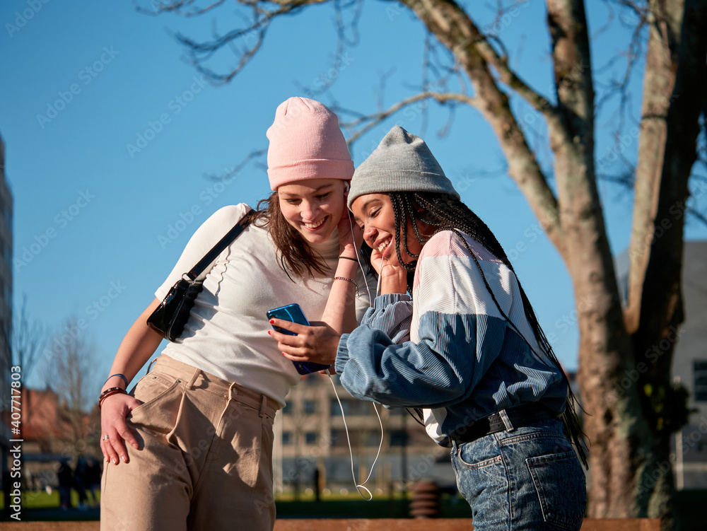 two young girl friends sharing a mobile phone outdoors, looking cheerful and wearing casual clothes