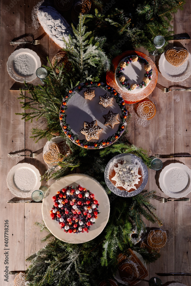 Decorated festive wooden table with white cake, cupcake, pie. Christmas decor of berries, thuja branches and a Christmas tree, sprinkled with snow. View from above