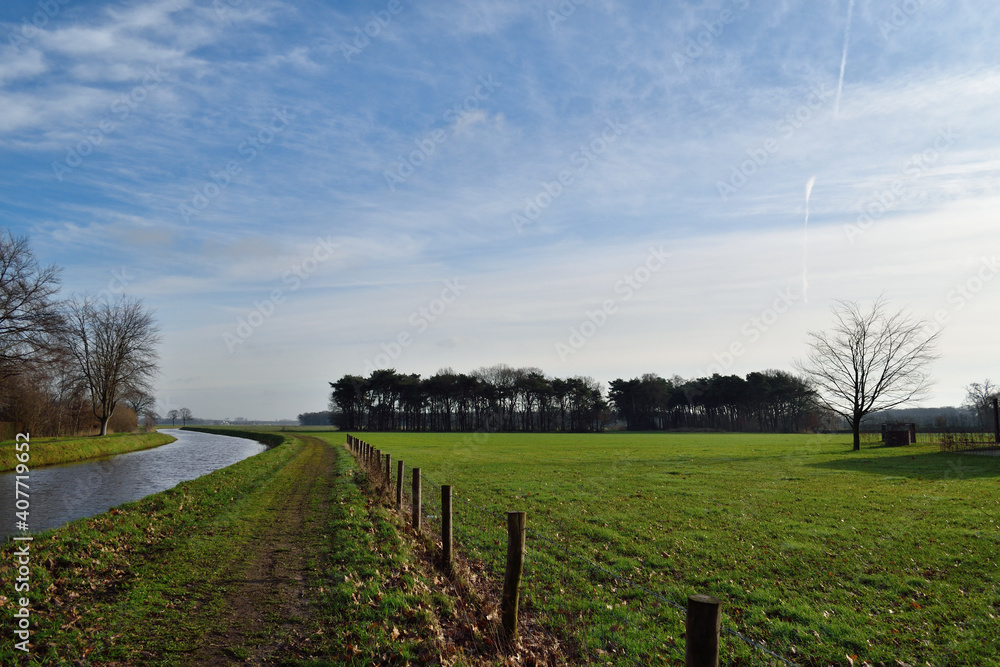 A landscape view of a Dutch canal with a footpath and fields beyond under a big, blue, winter sky 
