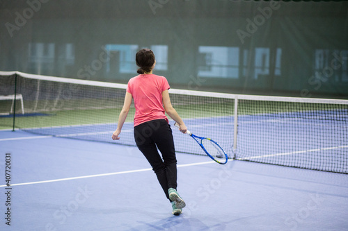 A young white brunette girl with a racket in her hands and in black sweatpants and a pink T-shirt is playing tennis and smiling. It is located in the indoor table tennis hall. © Dina