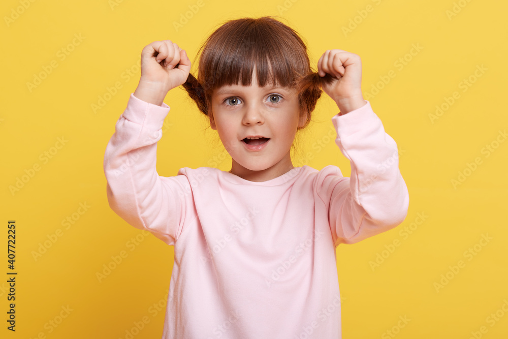 Happy little girl with pigtails looking directly at camera with opened mouth, keeping hands on her braids, wearing pale pink casual attire, posing isolated over yellow background. Happy little girl with pigtails looking directly at camera with opened mouth, keeping hands on her braids, wearing pale pink casual attire, posing isolated over yellow background.