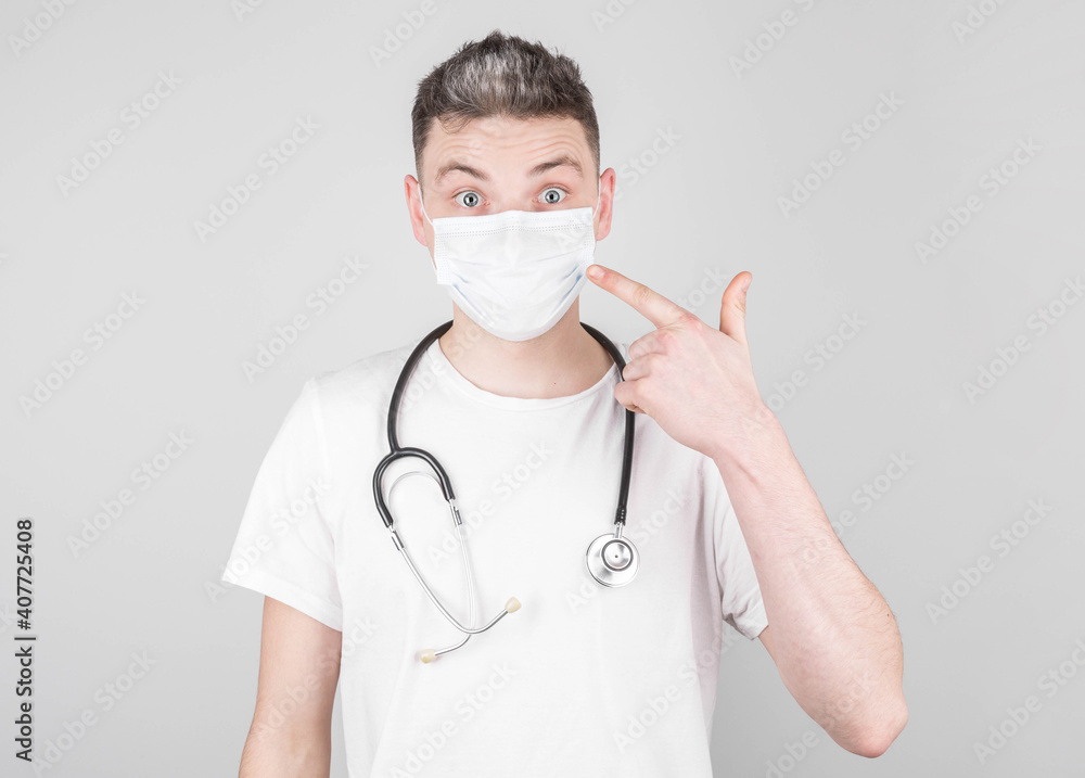 Male doctor in medical uniform points his fingers at a medical mask on his face. Concept of protection and prevention of covid-19, colds and flu