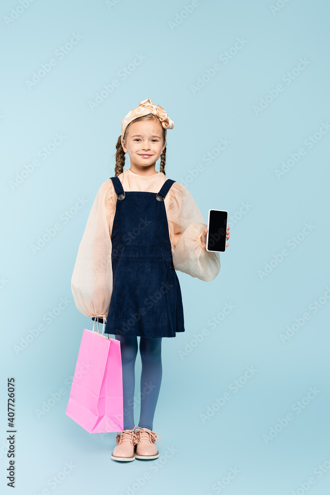 full length of happy kid with pigtails holding smartphone with blank screen and shopping bag on blue full length of happy kid with pigtails holding smartphone with blank screen and shopping bag on blue
