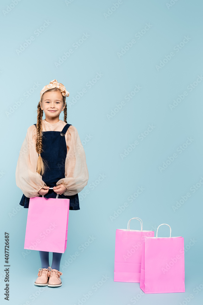 full length of little girl in dress smiling near shopping bags on blue full length of little girl in dress smiling near shopping bags on blue