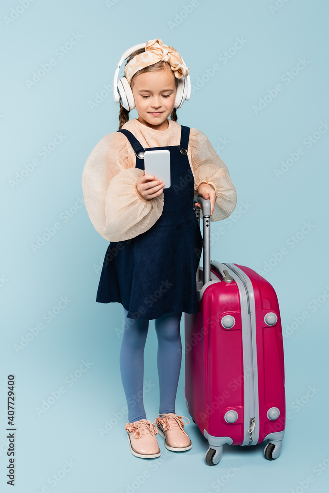 full length of little girl in wireless headphones using smartphone while standing with baggage on blue full length of little girl in wireless headphones using smartphone while standing with baggage on blue