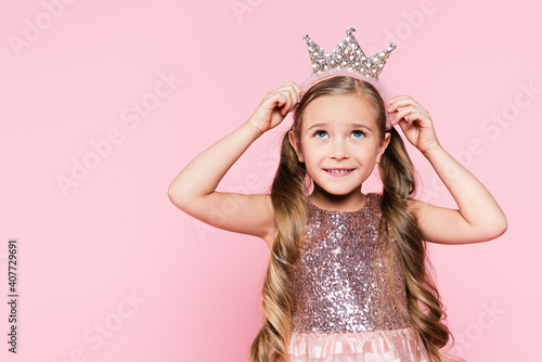 cheerful little girl in dress adjusting crown isolated on pink