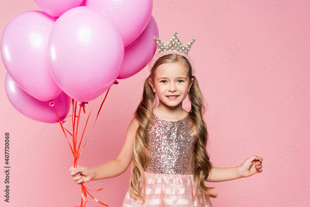cheerful little girl in dress and crown holding balloons isolated on pink