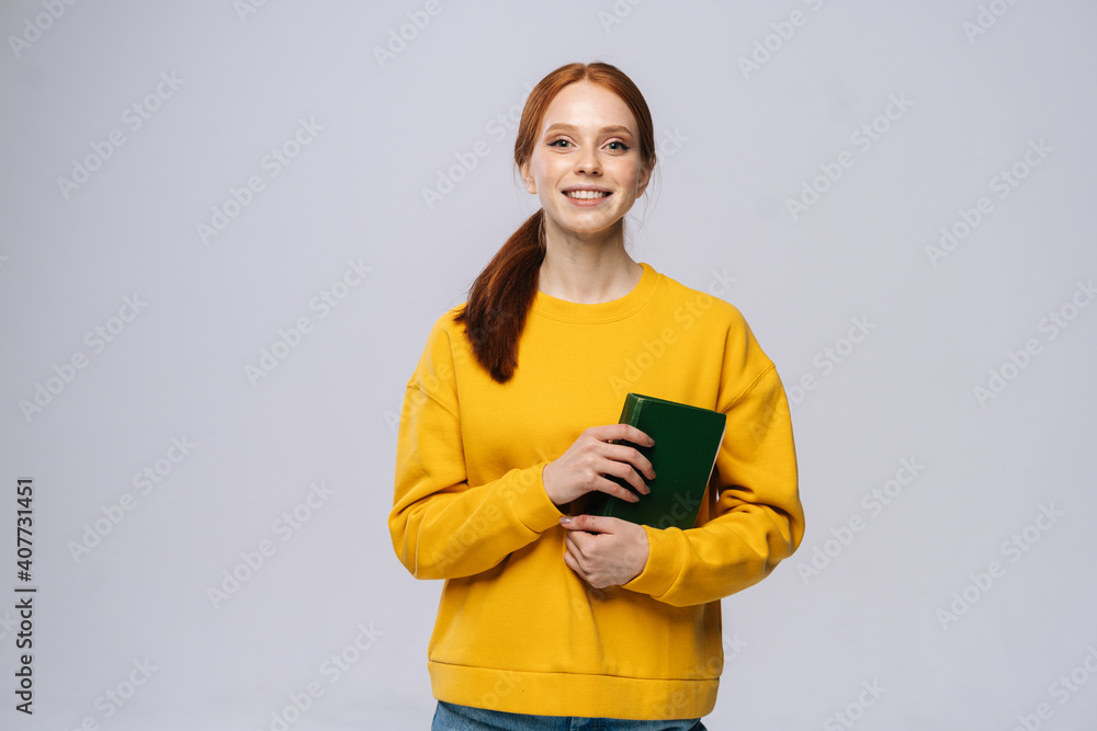 Smiling happy young woman college student yellow sweater holding book on isolated gray background. Pretty redhead lady model emotionally showing facial expressions, copy space. Smiling happy young woman college student yellow sweater holding book on isolated gray background. Pretty redhead lady model emotionally showing facial expressions, copy space.