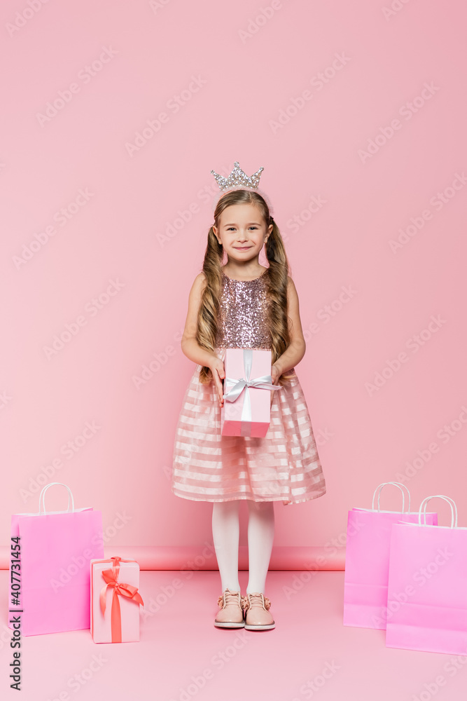 full length of smiling little girl in crown holding present near shopping bags on pink full length of smiling little girl in crown holding present near shopping bags on pink