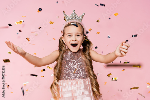 amazed little girl in crown with outstretched hands near falling confetti on pink