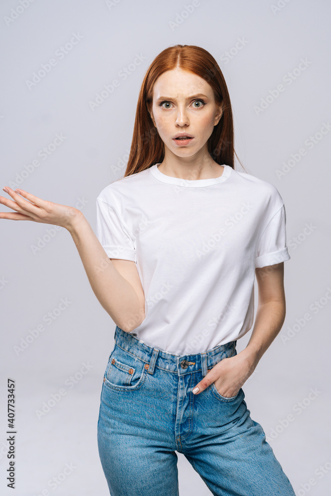 Shocked confused young woman wearing T-shirt and denim pants with opened mouth and raised hands on isolated white background. Pretty lady model with red hair emotionally showing facial expressions. Shocked confused young woman wearing T-shirt and denim pants with opened mouth and raised hands on isolated white background. Pretty lady model with red hair emotionally showing facial expressions.