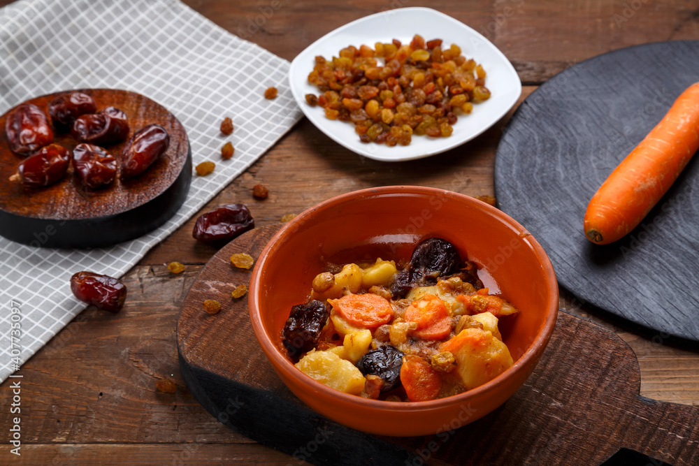A dish of Jewish cuisine sweet tsimes with carrots with dates in a plate next to a spoon and carrots with dried fruits. A dish of Jewish cuisine sweet tsimes with carrots with dates in a plate next to a spoon and carrots with dried fruits.