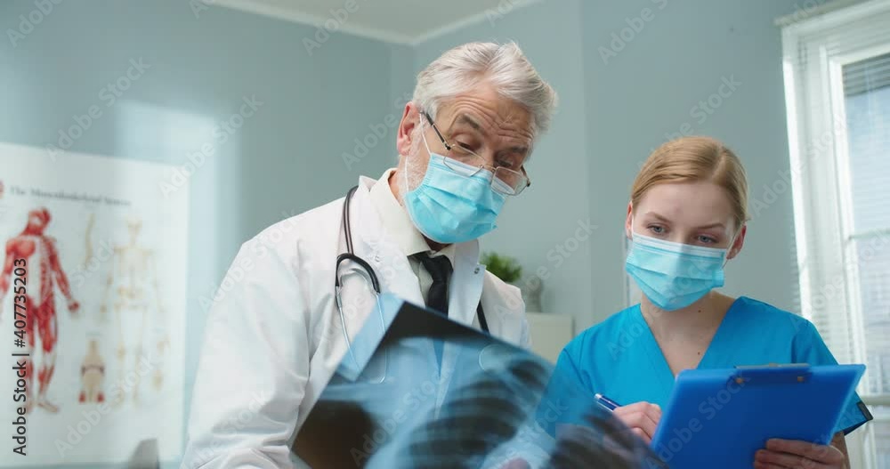 Caucasian medical professionals doing report of patient's disease in hospital office. Good-looking female nurse writing down data and talking to senior male doctor holding x-ray scan. Health. Caucasian medical professionals doing report of patient's disease in hospital office. Good-looking female nurse writing down data and talking to senior male doctor holding x-ray scan. Health.