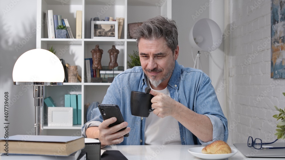 50s man having breakfast checking news on phone drinking morning coffee at home. Confident happy smiling.