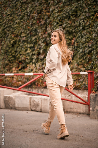 A pretty blonde young woman is walking down the street, she is wearing jeans and a beige shirt. Beautiful girl dressed in casual style with a smile on her face for a walk